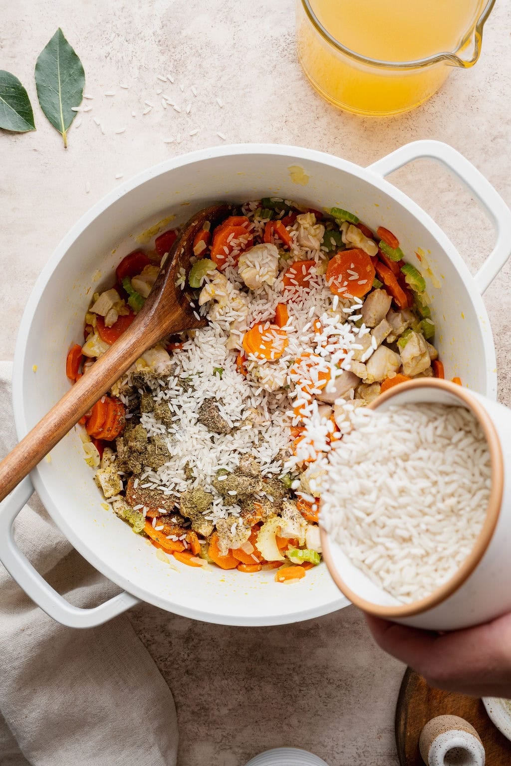 White rice being poured into a pot of seasoned carrots, celery, onion, and chicken.